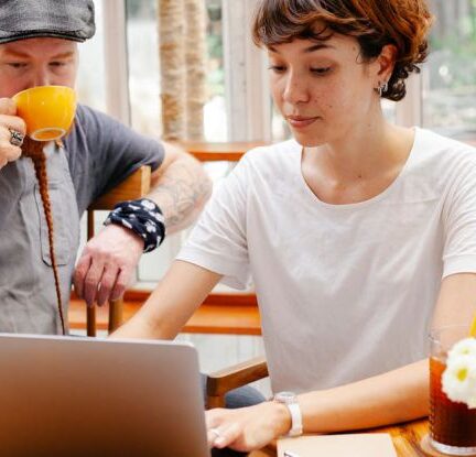 Remote Teams - Pensive young woman typing on netbook while man with cup drinking coffee at table of cafe